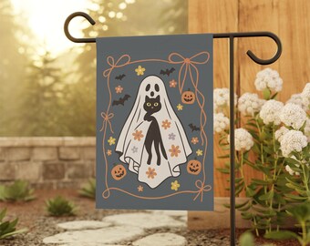 May include: A rectangular garden flag with a dark gray background featuring a central illustration of a black cat wearing a white ghost sheet. The design includes orange jack-o'-lanterns, small bats, and colorful flowers framed by an orange border.