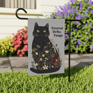 Può includere: Un gatto nero con gli occhi gialli è circondato da fiori colorati. Il gatto è seduto su uno sfondo grigio con il testo "Fox Trot Delta Tango" stampato sul lato destro.