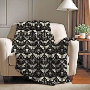 May include: A black throw blanket draped over a cream-colored armchair. The blanket features a repeating pattern of white moths, skulls, and ornate floral elements. The blanket adds a gothic or alternative aesthetic to the room.