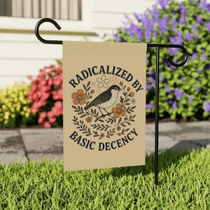 Peut inclure: Drapeau de jardin beige avec un support en métal noir. Il présente une illustration d'oiseau entourée de fleurs et le texte "RADICALIZED BY BASIC DECENCY" dans un design circulaire. Le drapeau est exposé dans un jardin avec des fleurs et de l'herbe.