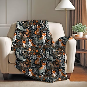 May include: A patterned throw blanket on a cream armchair. The blanket displays a repeating design of foxes, foliage, and flowers in orange, grey, and white against a dark blue backdrop. The blanket is made of a soft material, perfect for adding warmth to a room.