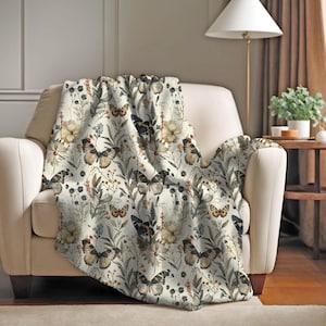 May include: A cream-colored throw blanket draped over a beige armchair. The blanket features a repeating pattern of butterflies, flowers, and foliage in shades of brown, beige, and green. The overall theme is nature and home decor.
