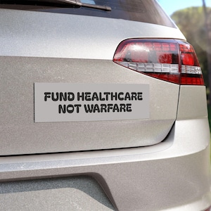 Puede incluir: Una pegatina rectangular con el texto "FUND HEALTHCARE NOT WARFARE" en negro, centrada en un fondo gris claro. La pegatina está adherida a la parte trasera de un coche plateado, cerca del piloto trasero.