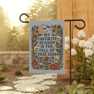 May include: A gray garden flag with the text "MY FAVORITE SEASON IS THE FALL OF FASCISM" surrounded by orange flowers, pumpkins, skulls, and a crescent moon with bats. The flag is displayed on a black metal stand.
