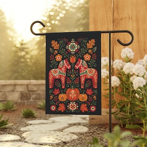 May include: A black garden flag featuring a folk art design with two red horses facing each other. The design includes pumpkins, flowers, and leaves in orange, green, and red. The flag is displayed on a black metal stand.