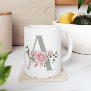 Puede incluir: Taza de cerámica blanca con la letra "A" en verde apagado, adornada con rosas rosas y hojas verdes. La taza está sobre un posavasos beige. Un artículo decorativo.