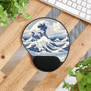 Może przedstawiać: Okrągła, ergonomiczna podkładka pod mysz z nadrukiem "Wielkiej Fali w Kanagawie" Hokusai. Projekt przedstawia dużą, stylizowaną falę w odcieniach niebieskiego i białego, z czarnym podparciem nadgarstka. Na drewnianej powierzchni.
