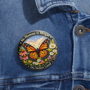 May include: A round button with a monarch butterfly design, surrounded by flowers and the text "No Human is Illegal" and "On Stolen Land". The butterfly has orange and black wings, set against a sky background.