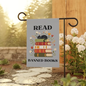 May include: A grey garden flag displaying the text "READ BANNED BOOKS". The flag features a stack of books with a black cat resting on top, surrounded by floral illustrations. The flag is mounted on a black metal stand.
