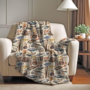 May include: A cream-colored throw blanket with a repeating pattern of watercolor illustrations of fishing-related items, including fish, fishing rods, lures, and tackle boxes.