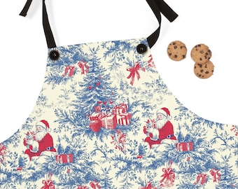 May include: A cream-colored apron with a blue and red Christmas toile pattern featuring Santa Claus, gifts, and Christmas trees. The apron has black ties and two black buttons. Three chocolate chip cookies are next to the apron.