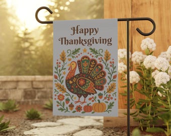 Può includere: Una bandiera da giardino con la scritta "Happy Thanksgiving" in lettere marroni. La bandiera presenta un tacchino colorato circondato da foglie autunnali, zucche e fiori. La bandiera è esposta su un supporto in metallo nero in un giardino.
