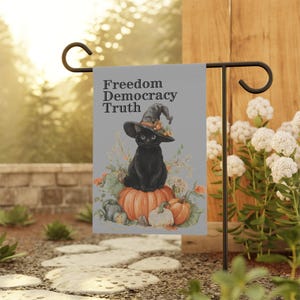 Può includere: Una bandiera da giardino con un gatto nero con un cappello da strega, seduto su una zucca. La bandiera ha le parole "Freedom Democracy Truth". La bandiera è su un supporto di metallo nero in un giardino.