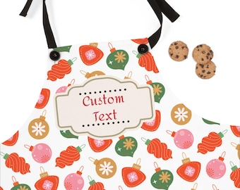 Op de afbeelding: Witte schort met een feestelijk kerstornamentpatroon in rood, groen, roze en goud. De schort heeft een aanpasbaar label met de tekst "Custom Text". Zwarte bandjes en een paar chocolate chip cookies zijn ook zichtbaar.
