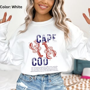 Puede incluir: Sudadera blanca con las palabras "CAPE COD" en azul y dos ilustraciones de langostas rojas. La sudadera tiene cuello redondo y mangas largas. El texto "Color: White" está en la esquina superior izquierda.