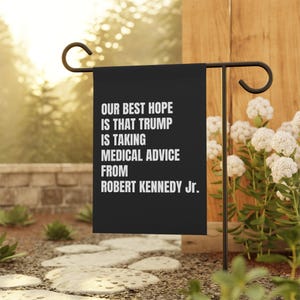 May include: Black garden flag with white text: "OUR BEST HOPE IS THAT TRUMP IS TAKING MEDICAL ADVICE FROM ROBERT KENNEDY Jr." The flag is displayed outdoors on a black metal stand, surrounded by greenery and flowers.