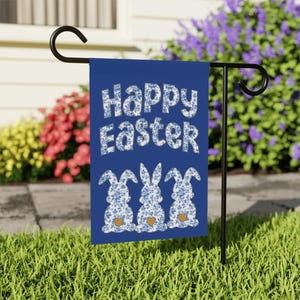 May include: A blue Easter garden flag with the words "Happy Easter" in white floral print. Three bunny silhouettes with floral patterns and orange hearts are below the text. The flag is displayed on a black metal stand in a garden.