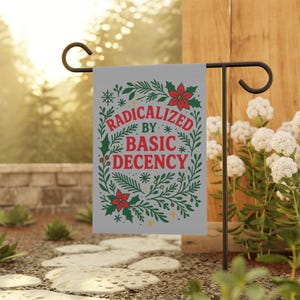 Pode incluir: Uma bandeira de jardim cinza com o texto "RADICALIZED BY BASIC DECENCY" em um design floral decorativo verde e vermelho. A bandeira é exibida em um suporte de metal preto em um jardim.