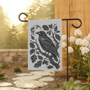 May include: A rectangular garden flag featuring a black and white illustration of a raven perched on an oak branch with acorns and a full moon in the background. The flag is suspended from a black metal stand, displaying a detailed nature scene.
