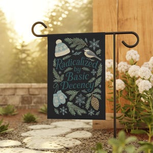 May include: Black garden flag with the text "Radicalized by Basic Decency" in teal script. The flag features illustrations of a winter hat, mittens, a bird, snowflakes, and pine needles. The flag is displayed on a black metal stand.