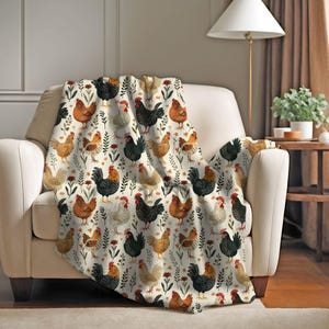 May include: A cream-coloured throw blanket with a repeating pattern of chickens in brown, black, and white, with floral accents. The blanket is draped over a chair and appears to be made of a soft material.