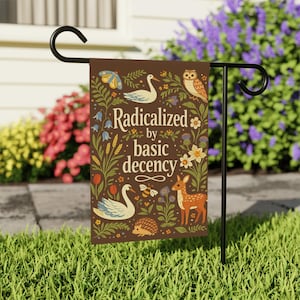 Pode incluir: Uma bandeira de jardim castanha com o texto "Radicalized by basic decency", rodeada por ilustrações de um cisne, uma coruja, um veado, uma abelha, um ouriço e flores. A bandeira está num suporte de metal preto num jardim.