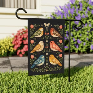May include: A black garden flag featuring a symmetrical design of colorful birds, flowers, and a butterfly. The birds are in shades of green, orange, yellow, and blue, set against a backdrop of green foliage and pink flowers. The flag is displayed on a black metal stand.