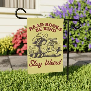 Puede incluir: Una bandera de jardín amarilla con un mapache con sombrero de vaquero leyendo un libro. El texto "Read Books Be Kind Stay Weird" está en letras granates. La bandera se muestra en un soporte de metal negro en un jardín cubierto de hierba.