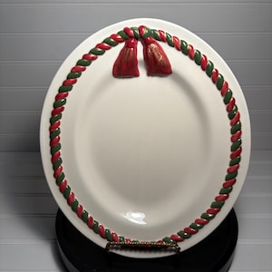 May include: A white ceramic plate with a red and green braided border and two red tassel accents. The plate is displayed on a black stand, showcasing a festive, holiday-themed design.