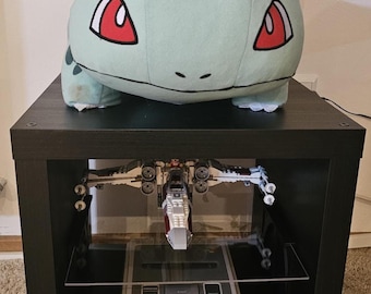 Puede incluir: Una criatura de peluche de color verde claro con un bulbo verde grande en la espalda, colocada sobre un estante cuadrado negro. Dentro del estante hay un modelo de caza estelar blanco y rojo sobre una consola Super Nintendo Entertainment System.
