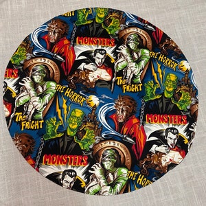 May include: A round fabric coaster with a colorful print of various monsters. The print features a variety of monsters, including Frankenstein, Dracula, and a werewolf. The text "The Horror", "The Fright", and "Monsters" is repeated throughout the print.