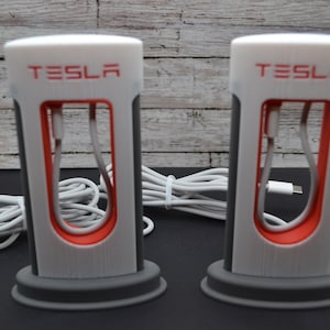 May include: Two white and grey Tesla charging station stands with red accents and the word "TESLA" printed on the front. Each stand has a white cable coiled inside.