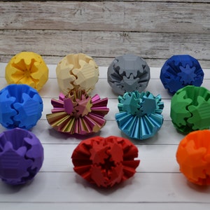 May include: A collection of 12 colourful 3D printed gear balls. The balls are arranged in a circle on a white surface. The balls are made of plastic and have a variety of colours, including red, orange, yellow, green, blue, purple, and grey.