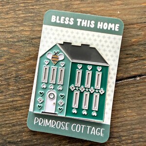 May include: A teal and green house-shaped enamel pin with a black roof, a bee, and the text "BLESS THIS HOME" and "PRIMROSE COTTAGE". The pin has white windows and a white door, with heart and flower details.