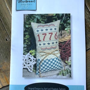 May include: A printed design featuring a decorative pillow with the number "1776" in red. The pillow has a blue and white checkered bottom, a beige center with red embroidery, and is tied with twine and lace. The design is on a white background.