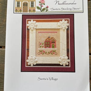 May include: A cross-stitch pattern featuring a red and green house with a green door, surrounded by a white frame with floral accents. The text reads "Country Cottage Needleworks" and "Santa's Stocking Store."