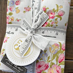 May include: A bundle of floral fabric tied with a white ribbon printed with a ruler design. The fabric features pink, yellow, and blue flowers on a light background. A round tag with gold text and a black tag with white text are attached.