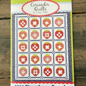 May include: A quilt pattern titled "Coriander Quilts by Corey Yoder" featuring a strawberry sampler design. The pattern showcases a grid of red strawberry blocks with various geometric patterns on a white background, framed by a blue border.