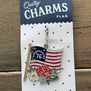 May include: A patriotic flag charm with red and white stripes, a blue field with stars, and the number 76. The charm is adorned with floral accents and attached to a silver clasp. The packaging reads "Quilty CHARMS FLAG".