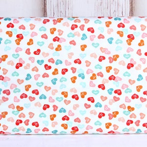 May include: White fabric with a repeating pattern of small, colorful hearts. Each heart has a different color and a white inscription inside. The colors of the hearts are red, orange, pink, blue, and teal.