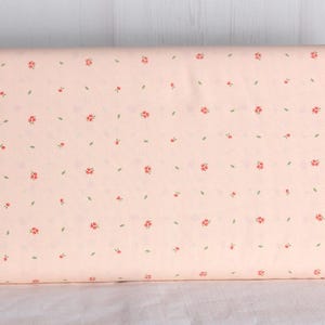 May include: A roll of fabric with a light peach background. The fabric is patterned with small red flowers and green leaves. The floral pattern is evenly spaced across the fabric.