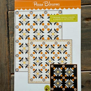 May include: A quilt pattern titled "Pecan Blossoms" with a repeating geometric design in orange, black, and cream. The pattern includes the text "CHOOSE FROM 3 SIZES & DESIGN OPTIONS!" and the logo of "FIG TREE & Co."