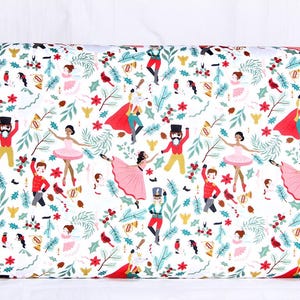 May include: A white fabric featuring a repeating pattern of nutcrackers in red and gold uniforms, ballerinas in pink dresses, red cardinals, green pine branches, and red holly berries. The design includes small gold trumpets and pinecones.