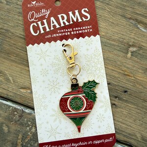 May include: A metal Christmas ornament charm with a gold-tone lobster clasp, featuring red, green, and white enamel details in a teardrop shape with holly leaves. The charm is attached to a branded Riley Blake Designs Quilty Charms card.