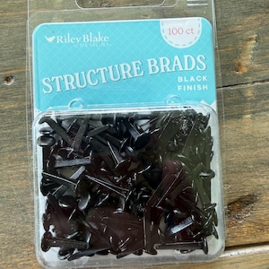 May include: A clear plastic package containing 100 black metal structure brads by Riley Blake Designs. The packaging features a light blue label with white text that reads Structure Brads, Black Finish, and 100 ct, showcasing the fasteners.
