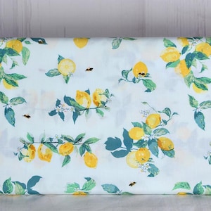 May include: White fabric with a repeating pattern of yellow lemons, green leaves, and small black bees. The lemons are in various stages of ripeness, some whole and some sliced. The leaves are a mix of green and teal. A cheerful, summery design.