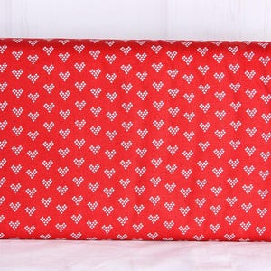 May include: Red fabric with a repeating pattern of white pixelated hearts. The fabric is folded, showing the pattern across the surface. The material appears to be cotton or a similar textile, suitable for various crafting projects.