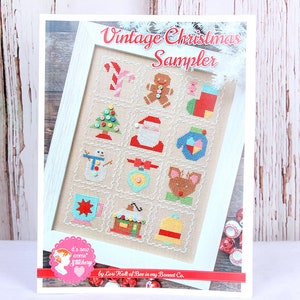 May include: A framed cross-stitch sampler titled "Vintage Christmas Sampler." The sampler features various Christmas-themed designs, including a candy cane, gingerbread man, Christmas tree, Santa Claus, snowman, ornament, and reindeer. The frame is white.