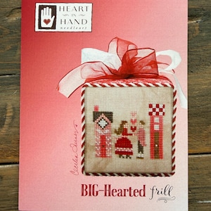 May include: A cross-stitch kit with a red and white ribbon and bow. The kit features a cross-stitched design of a house, people, and a tower in shades of red and white. The text "BIG-Hearted frill" is at the bottom, and the Heart in Hand Needleart logo is at the top.