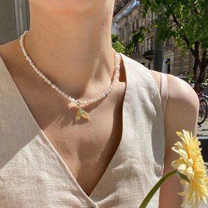 May include: A woman wearing a beige linen dress with a pearl necklace and a small gold pendant. She is holding a yellow flower.